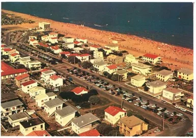 Vue aérienne de Marseillan plage avec des maisons blanches aux toits rouges, une plage de sable doré en arrière-plan et des voitures garées le long de la route principale. De nombreuses personnes profitent de la plage et de l'eau bleue en bord de mer, créant une ambiance estivale animée.